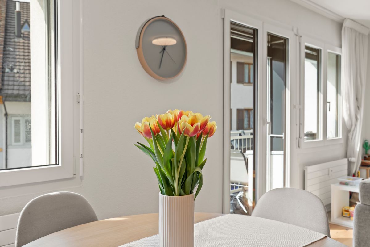 Essbereich mit Tisch, Blumen und großen Fenstern.