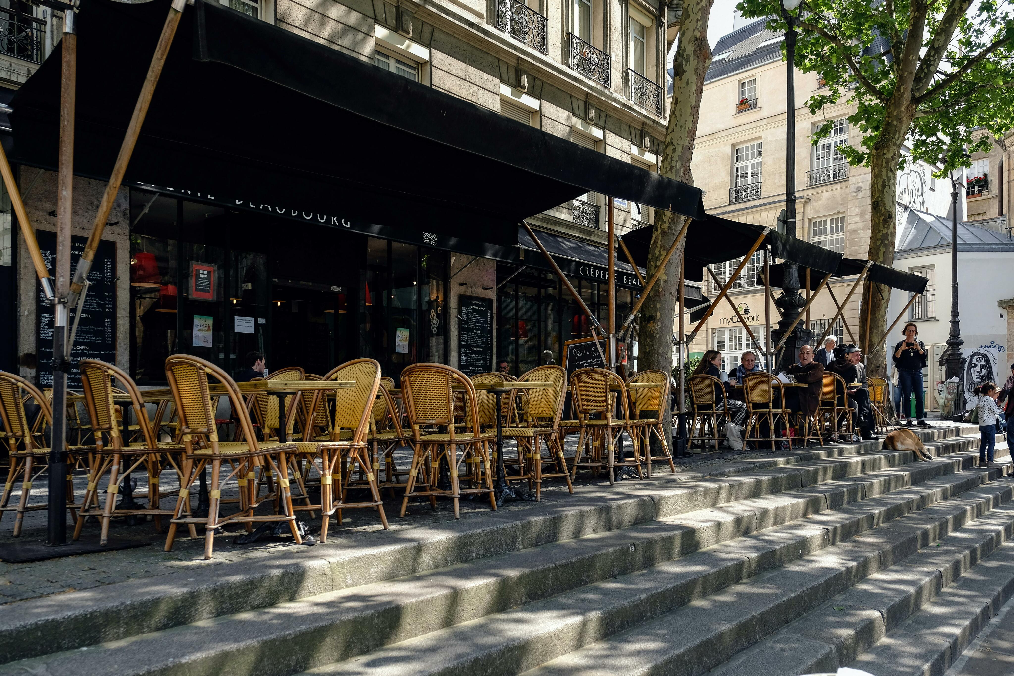 un trottoir avec des marches, des tables et des chaises avec des parasols et des gens assis sous un arbre