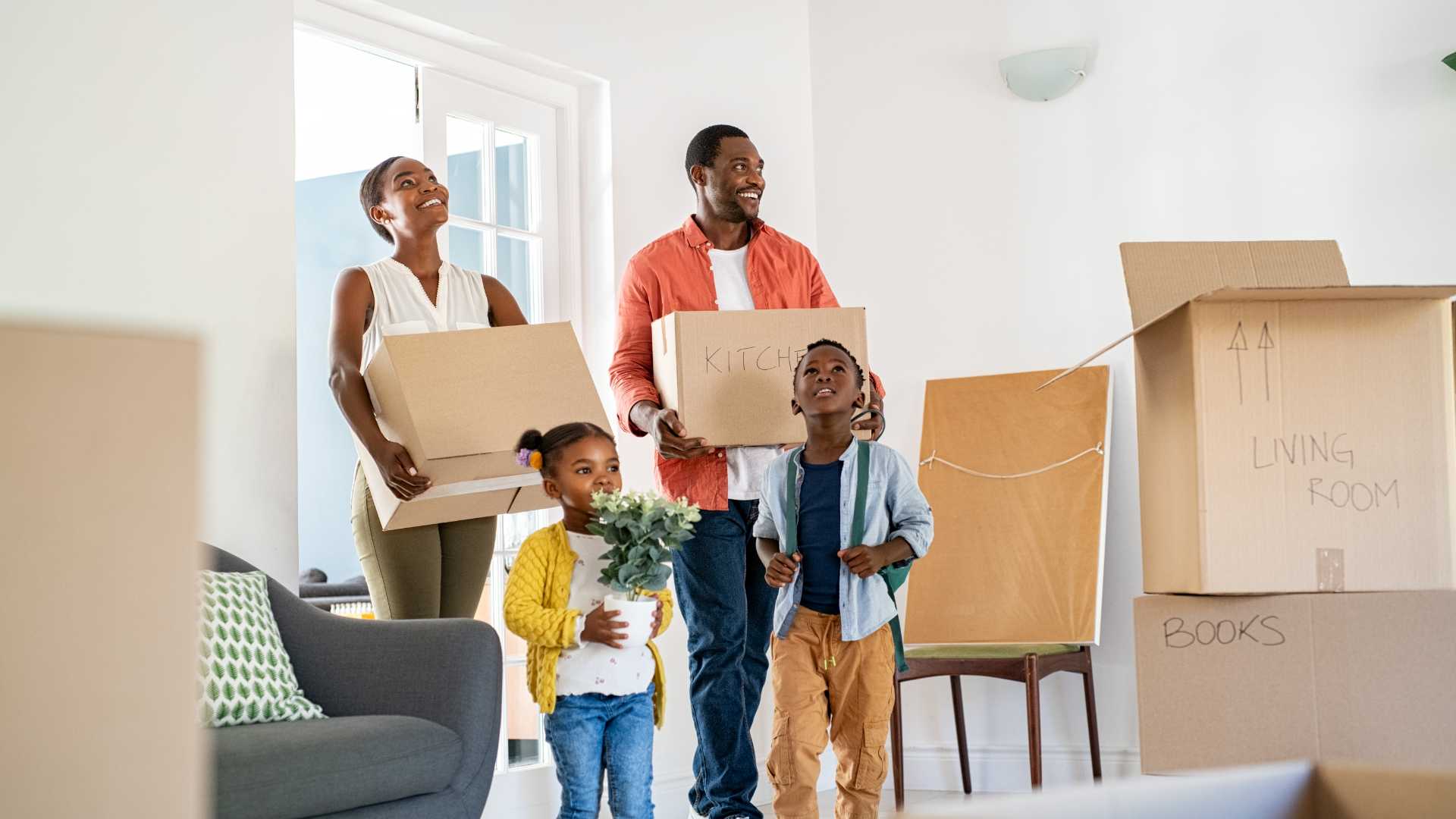 Eine Familie mit zwei Kindern betritt eine helle Wohnung und trägt Umzugskartons, die mit Wohnräumen beschriftet sind.