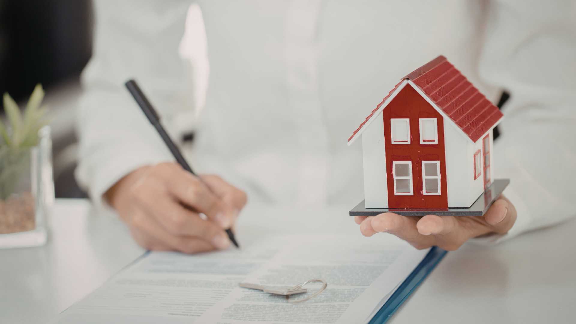 Une personne tient une petite maison dans sa main tout en signant un contrat.