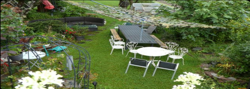 une petite cour avec une pelouse verte et beaucoup de plantes, une table et des chaises blanches une petite cour avec une pelouse verte et beaucoup de plantes, une table et des chaises blanches