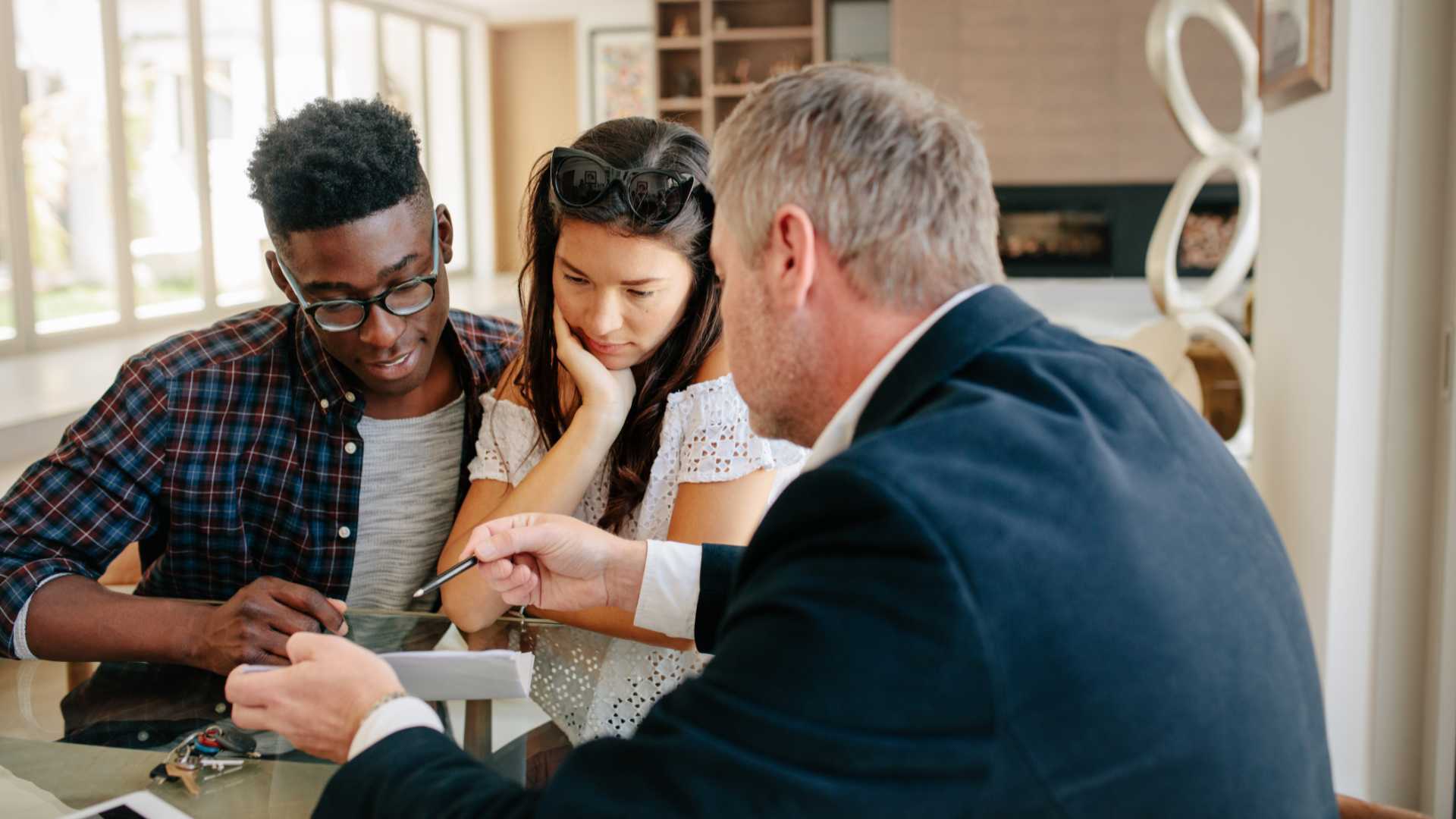 Una coppia consulta un consulente bancario in un ufficio luminoso, esaminando documenti finanziari e opzioni di mutuo per diventare proprietari di casa.